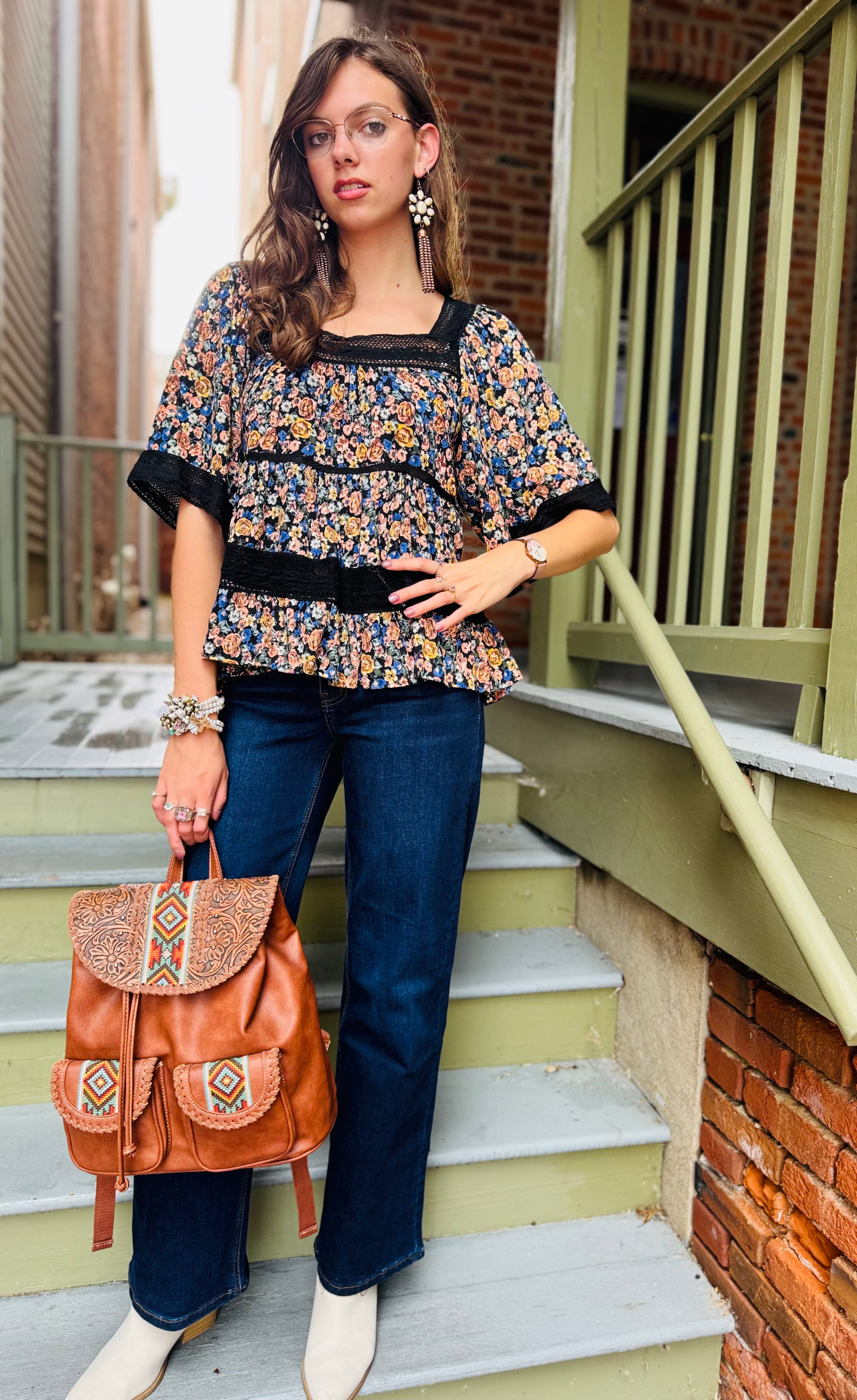 Woman standing on outdoor steps wearing a floral top and blue jeans, holding a brown leather bag.